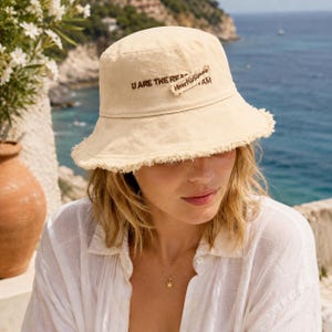 Peut inclure: Un chapeau bob beige avec un bord effiloché et un texte brodé en brun foncé indiquant U ARE THE REASON et Heart of Summer. Le chapeau présente une calotte structurée et un large bord incliné vers le bas, fabriqué dans un tissu uni clair.