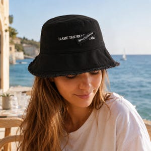 Peut inclure: Un chapeau bob noir avec un bord effiloché et un texte brodé en blanc indiquant U ARE THE REASON et Heart is simple I AM. Le chapeau présente une calotte structurée et un bord incliné vers le bas, fabriqué dans un tissu noir uni.