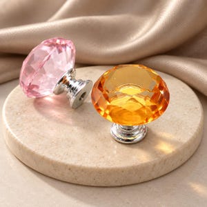Puede incluir: Dos pomos para armario facetados con forma de diamante. Un pomo es de color rosa translúcido y el otro es de color ámbar translúcido. Ambos cuentan con una base de metal plateado pulido con un punto de montaje circular y un orificio roscado.