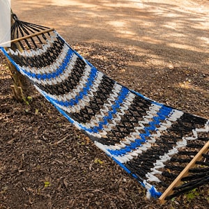 May include: A woven hammock featuring a repeating striped pattern in black, white, and blue. The hammock is suspended between two wooden spreader bars at each end, with black rope attachments extending from the bars to secure the structure.
