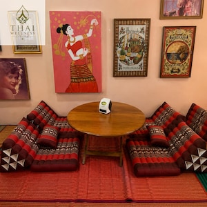 May include: A round wooden table sits between two red and dark brown Thai floor cushions with triangular backrests. The cushions feature a repeating elephant pattern. A small white air purifier is placed on the center of the wooden table.