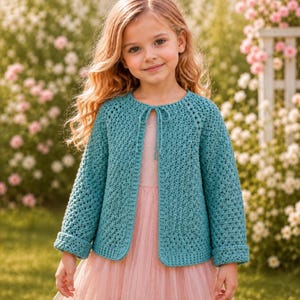 May include: A teal long-sleeved crochet cardigan featuring a textured stitch pattern, a round neckline, and a thin tie closure at the front. The garment is worn over a light pink dress, showcasing a consistent knit construction throughout the sleeves.