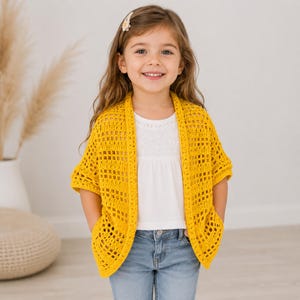 Può includere: Un cardigan all'uncinetto giallo brillante aperto sul davanti, caratterizzato da un motivo a griglia quadrata, maniche corte e due tasche frontali. Il capo è indossato sopra un top bianco a maniche corte e jeans blu, con una trama lavorata a maglia.