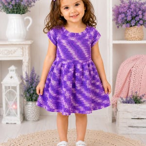 May include: A short-sleeved crochet dress featuring a vibrant purple and white variegated yarn pattern. The garment has a round neckline, a fitted bodice, and a flared skirt that falls above the knees, showcasing a consistent textured stitch.