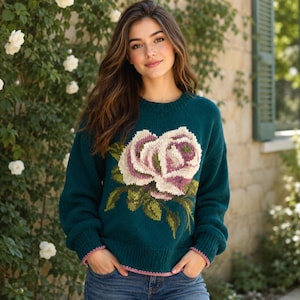 May include: A teal long-sleeve knit sweater featuring a large, central intarsia rose design in shades of cream, light pink, and dark pink, with green leaves. The sweater has a ribbed crew neckline, cuffs, and hem with a thin pink trim detail.