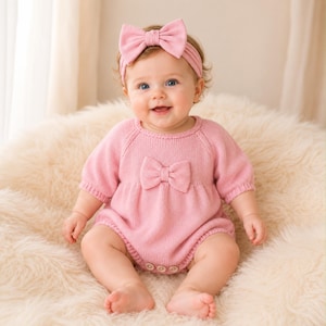 May include: A light pink knitted baby romper featuring short sleeves, a round neckline, and a central bow detail on the chest. The garment includes three small buttons at the bottom closure. A matching light pink headband with a large bow is worn.