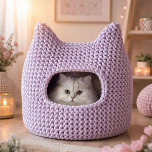 May include: A light purple, chunky knit cat bed shaped like a cat head with two pointed ears. The bed features a large, oval-shaped opening in the center, revealing a fluffy, long-haired white cat with green eyes resting inside the structure.
