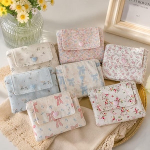 Puede incluir: Seis carteras pequeñas de tela con cierre de solapa, cada una con un patrón floral o de lazo único en colores pastel. Cada cartera está asegurada con un pequeño botón en forma de corazón. Los diseños incluyen estampados florales en azul claro, rosa y crema.