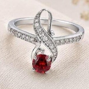 Può includere: Un anello color argento con una pietra preziosa rossa dal taglio rotondo al centro e un design a fascia avvolgente, arricchito da piccole pietre chiare lungo il gambo e l'elemento centrale curvo, dall'aspetto metallico lucido.
