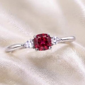 Peut inclure: Une bague argentée ornée d'une pierre précieuse centrale rouge foncé taille coussin, encadrée par deux pierres transparentes plus petites en forme de poire. L'anneau est lisse et poli, présentant un design à trois pierres avec un centre rouge.