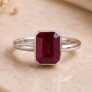 Op de afbeelding: Een zilveren ring met een rechthoekige, dieprode edelsteen in emerald-slijpvorm, gezet in een gladde zetting. De ringband is van gepolijst zilver met een afgerond profiel, wat zorgt voor een strak ontwerp dat de levendige rode steen accentueert.
