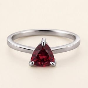 Op de afbeelding: Een zilverkleurige ring met een enkele dieprode edelsteen in een trilliant-slijpvorm, gezet in een zetting met drie pootjes. De ringband is glad en gepolijst, met een strak ontwerp dat de driehoekige vorm en de levendige kleur van de steen benadrukt.