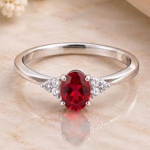 Könnte beinhalten: Ein silberfarbener Ring mit einem zentralen, oval geschliffenen roten Edelstein, der von vier Krappen gehalten wird. Die Ringschiene ist auf beiden Seiten mit je drei kleinen, klaren, rund geschliffenen Steinen in Dreiecksform besetzt.