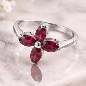May include: A silver-toned ring featuring a cluster of four deep red, marquise-cut gemstones arranged in a flower-like pattern. The band is smooth and polished, holding the stones in a central setting that creates a distinct floral silhouette.