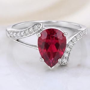 Op de afbeelding: Een zilverkleurige ring met een centrale peervormige, dieprode edelsteen die door vier pootjes wordt vastgehouden. De ringband is verdeeld in twee asymmetrische, gebogen armen die aan de bovenzijde zijn bezet met kleine, heldere steentjes.