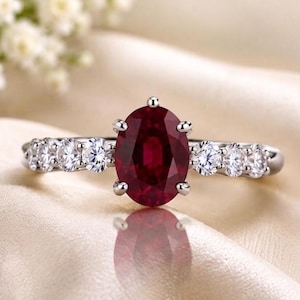 Può includere: Un anello color argento con una gemma rosso scuro dal taglio ovale, fissata da quattro griffe. Il gambo dell'anello è impreziosito da tre piccole pietre rotonde, trasparenti e dal taglio brillante su ciascun lato della montatura.