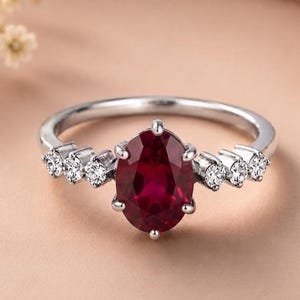 Può includere: Un anello color argento con una pietra preziosa rosso scuro dal taglio ovale fissata da sei griffe. Il gambo dell'anello è decorato su entrambi i lati da tre piccole pietre rotonde, trasparenti e sfaccettate, creando un design simmetrico.