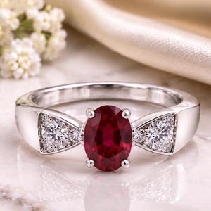 Könnte beinhalten: Ein silberfarbener Ring mit einem zentralen, oval geschliffenen, tiefroten Rubin, der von vier Krappen gehalten wird. Die Ringschiene hat zwei dreieckige Seitenteile, die jeweils mit drei kleinen, runden, klaren weißen Diamanten besetzt sind.