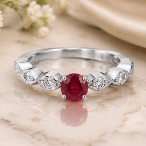 Könnte beinhalten: Ein silberfarbener Ring mit einem zentralen, rund geschliffenen, leuchtend roten Edelstein, der von vier Krappen gehalten wird. Die Ringschiene ist mit kleineren, rund geschliffenen, klaren Steinen in geometrischen, rautenförmigen Fassungen besetzt.