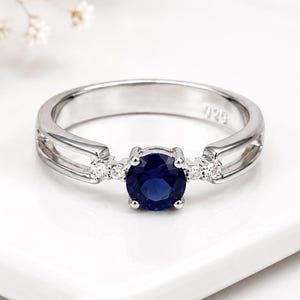 May include: A silver ring featuring a round, deep blue centre stone held by four prongs. The band is split on both sides, with two small, clear, round-cut stones set into each side of the shank. The interior of the band is stamped with 925.