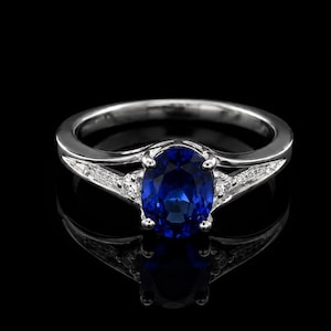 May include: A silver-toned ring featuring a central oval-cut deep blue gemstone held by four prongs. The band is split at the shoulders and accented with small, clear, round-cut stones embedded along the sides, creating a refined appearance.