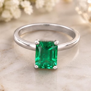 May include: A silver-toned ring featuring a single emerald-cut green gemstone set in a four-prong setting. The band is smooth and polished, showcasing a clean, metallic finish that complements the vibrant, rectangular green stone at the centre.