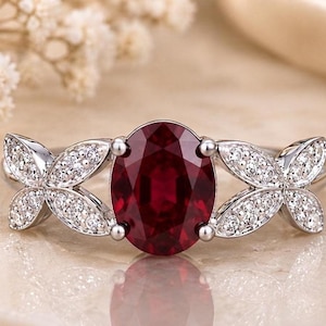 May include: A silver-toned ring featuring a large, oval-cut deep red gemstone at the center. The band is adorned on both sides with two leaf-shaped metal segments, each encrusted with small, clear, sparkling stones that catch the light.