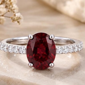 Pode incluir: Um anel de tom prateado com uma grande pedra preciosa de cor vermelho-escura e corte oval, presa por quatro garras. A banda é decorada com uma fila de pequenas pedras claras de corte redondo incrustadas em ambos os lados do aro do anel.