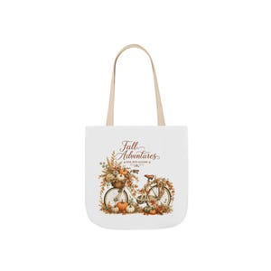 May include: A white tote bag with two beige handles featuring a graphic of a bicycle surrounded by autumn leaves and pumpkins. The text "Fall Adventures" and "RIDE INTO AUTUMN" is printed in brown script and serif font above the bicycle illustration.