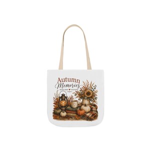 May include: A white tote bag with beige handles featuring a graphic of a lantern, a stack of books, a mug, pumpkins, and a sunflower bouquet. The text reads Autumn Memories MADE HERE together with a small heart icon, surrounded by fall foliage.