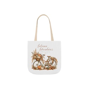 May include: A white tote bag with two beige handles featuring a graphic of a bicycle surrounded by autumn leaves and pumpkins. The text Autumn Adventures is printed in a brown script font above the bicycle, with smaller text underneath.