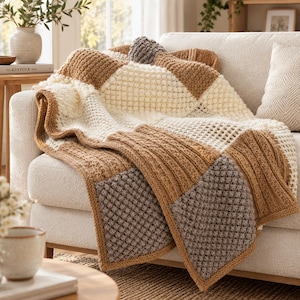 May include: A chunky knit throw blanket featuring a patchwork design of alternating squares in cream, tan, and grey. The blanket displays various textured stitch patterns, including bobble, ribbed, and waffle weaves, finished with a solid border.