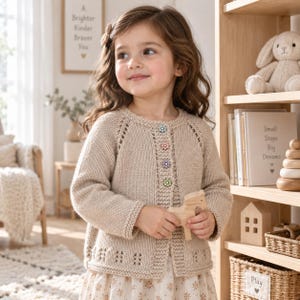 May include: A beige long-sleeved knit cardigan featuring a round neckline, a button-down front with four multi-coloured floral-shaped buttons, and a decorative open-work pattern along the hemline and raglan sleeve seams.