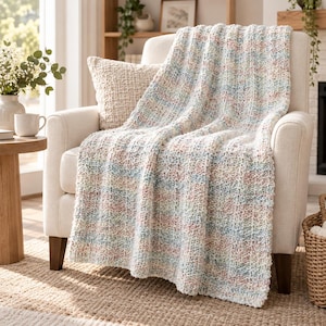 Peut inclure: Un plaid en tricot épais présentant un motif de fil panaché avec des taches pastel douces de bleu, de rose et de crème. La couverture a un design en maille côtelée texturée et est drapée sur un fauteuil rembourré de couleur crème.
