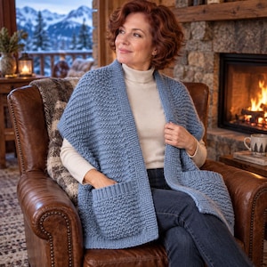 Può includere: Un cardigan in maglia senza maniche color azzurro con un motivo a maglia testurizzato, un ampio colletto a coste e due grandi tasche frontali applicate. Il capo è indossato sopra un top a maniche lunghe color crema e jeans blu scuro.