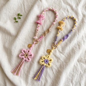 May include: Two macrame wristlets featuring braided cord loops in pastel pink, white, and tan, and yellow, purple, and white. Each wristlet includes a flower-shaped charm, a small gold-toned metal rabbit pendant, and a multi-colored cord tassel.