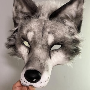 Può includere: Una maschera da lupo con una realistica texture in pelliccia sintetica grigia e bianca, orecchie a punta e naso nero. La maschera presenta aperture per gli occhi a mandorla e ombreggiature dettagliate attorno al muso e alla fronte.