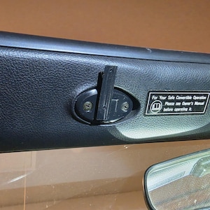 May include: A black plastic latch component installed on a textured black vehicle interior panel. The part features a vertical lever mechanism secured by two screws. A rectangular warning label with white text reads: For Your Safe Convertible Operation Please see Owner's Manual before operating it.