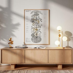 May include: A framed vertical wall art print featuring an exploded view diagram of a mechanical watch movement with gold and silver gears, labeled with technical text, displayed in a light wood frame on a light-colored wall above a wooden console.