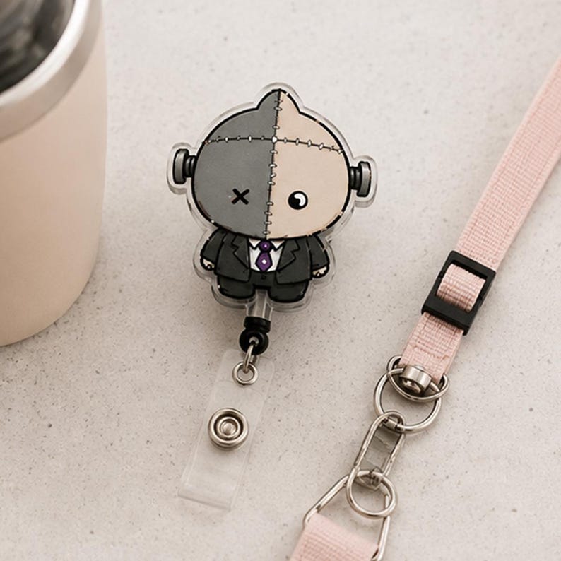 May include: A retractable badge reel featuring a split-colored character with a dark gray and beige head, wearing a black suit with a purple tie. The character has a stitched face, one X-shaped eye, and one round eye. It is attached to a light pink lanyard.