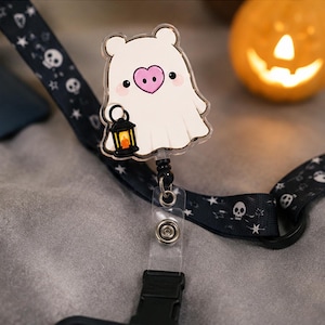 May include: A badge reel featuring a white ghost character with a pink heart-shaped nose holding a small black lantern. The ghost is attached to a black lanyard patterned with white skulls and stars, connected by a clear plastic strap and metal clip.