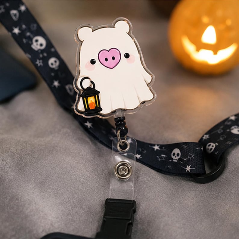 May include: A badge reel featuring a white ghost character with a pink heart-shaped nose holding a small black lantern. The ghost is attached to a black lanyard patterned with white skulls and stars, connected by a clear plastic strap and metal clip.