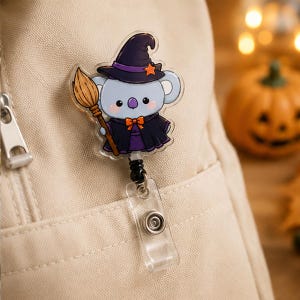 May include: A badge reel featuring a light blue koala wearing a dark purple witch hat with an orange star and a matching purple cape. The koala holds a brown broomstick. The badge reel has a clear plastic strap and a metal snap button attachment.