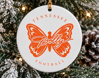 May include: A round white ceramic ornament featuring an orange butterfly graphic in the center. The text Tennessee is printed in an arch above the butterfly, the word Dolly is written in a script font across the butterfly, and Football is below.