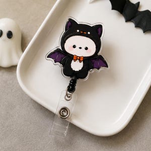May include: A retractable badge reel featuring a clear acrylic topper with a cartoon character wearing a black bat costume. The character has a white face, pink cheeks, and a small orange bow tie. The bat wings are purple and black.