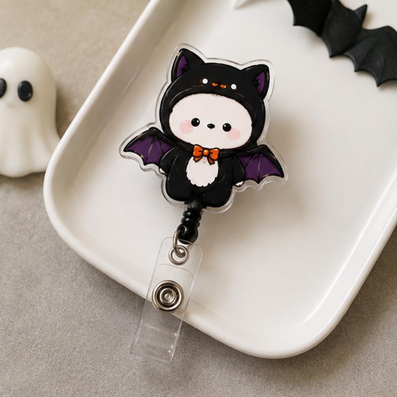 May include: A retractable badge reel featuring a clear acrylic topper with a cartoon character wearing a black bat costume. The character has a white face, pink cheeks, and a small orange bow tie. The bat wings are purple and black.