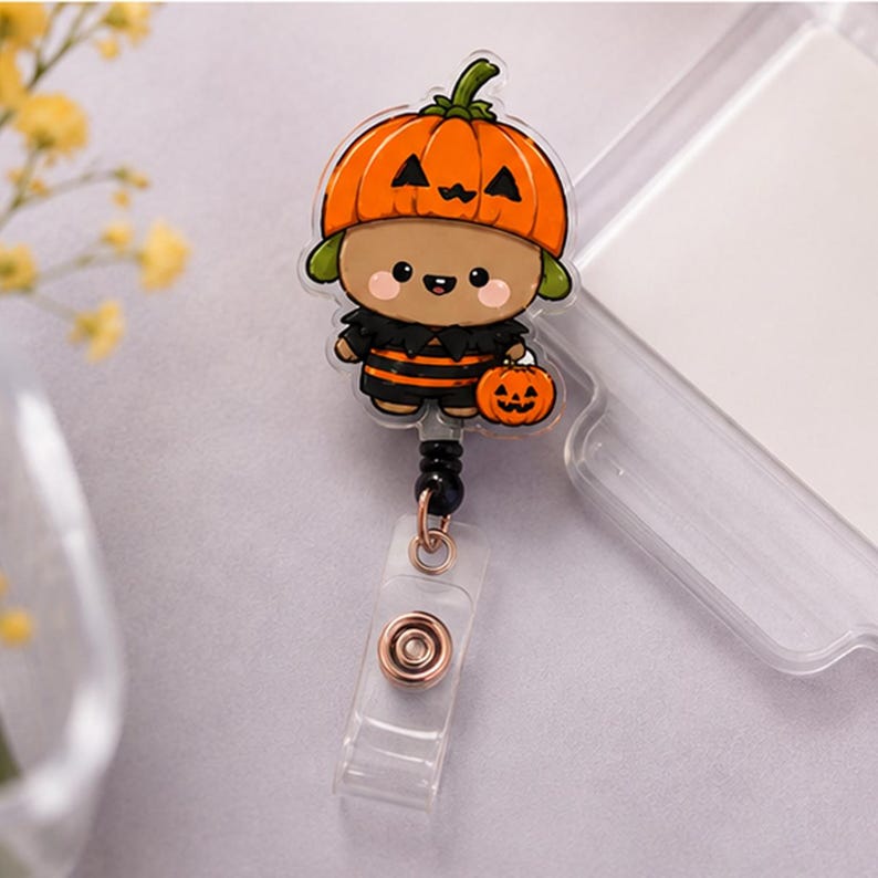 May include: A retractable badge reel featuring a cartoon character wearing an orange pumpkin hat and a black and orange striped outfit. The character holds a small jack-o'-lantern. The reel has a clear plastic strap with a metal snap button.
