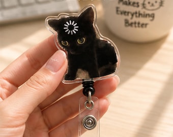 May include: A badge reel featuring a clear acrylic topper with a black cat graphic. The cat has a white loading icon on its forehead. The badge reel has a black retractable cord and a clear plastic strap with a metal snap button closure.