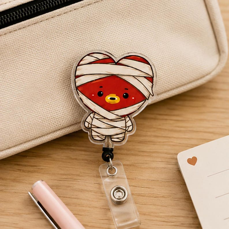 May include: A badge reel featuring a red heart-shaped character wrapped in white bandages like a mummy. The character has black eyes and a yellow mouth. The badge reel includes a clear plastic strap with a metal snap button for holding an ID card.