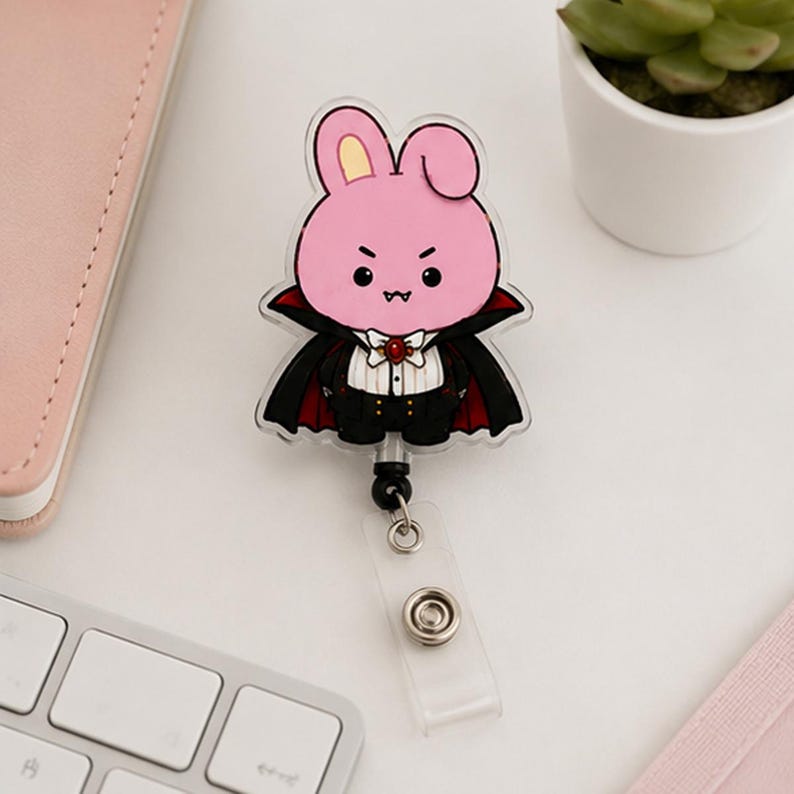 May include: An acrylic badge reel featuring a pink cartoon rabbit dressed as a vampire. The rabbit has a black cape with a red lining, a white shirt with a red gem brooch, and black trousers. The reel includes a clear plastic strap with a metal snap.